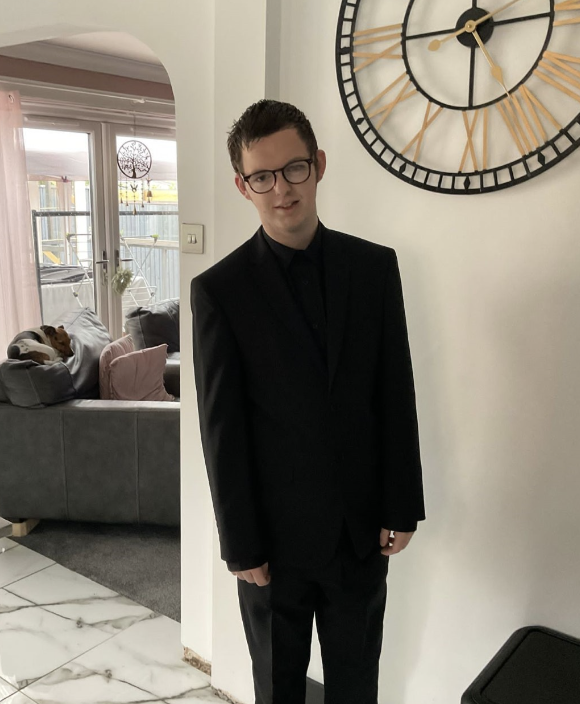 Zack, 19, is in a black suit and tie, looking excited to attend his prom. he has short hair, is white, and slim and wears glasses