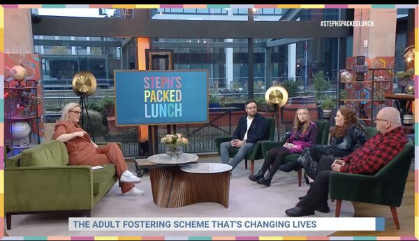 A freeze frame of pss' appearance on Steph's Packed Lunch. Steph sits on a coach to the left and the family sit on the right with a coffee table in between them and Steph. At the bottom of the screen reads a headline: adult fostering scheme changing lives