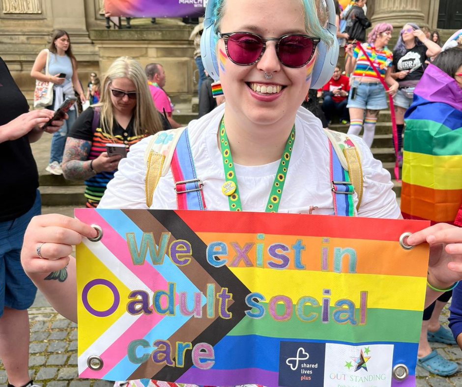 Hope has blue hair and wears purple tinted glasses. She has a huge smile and wears a white t shirt and multicoloured dungarees. She's holding up a handheld sign that has the LGBTQIA+ flag and reads 'we exist in adult social care' - her mental health is stronger than ever thanks to Shared Lives