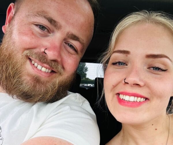Chloe and her partner are taking a selfie together. They are both white, one male, one female. Chlose is blond, youthful looking, and wearing pink lipstick. She has her nose pierced and has blue eyes. Her partner also has his nose pierced and has a full beard and moustache. He has blue eyes too. A caregiving job with a difference.