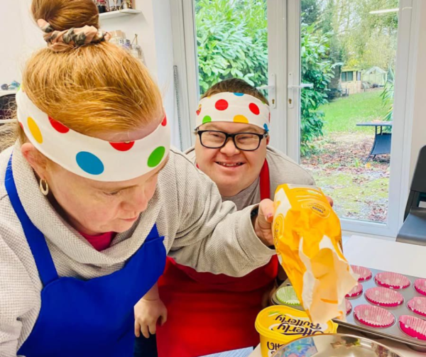 Victoria and someone who also uses Helena's day centre is cooking in the kitchen. They're wearing colourful polkadot headbands and aprons and Victoria is concentrating on weighing out flour. Her friend int he picture also has Down's Syndrome and smiling brightly at the camera. They're making cupcakes. Fostering adults can be as simple as baking a cake some days.